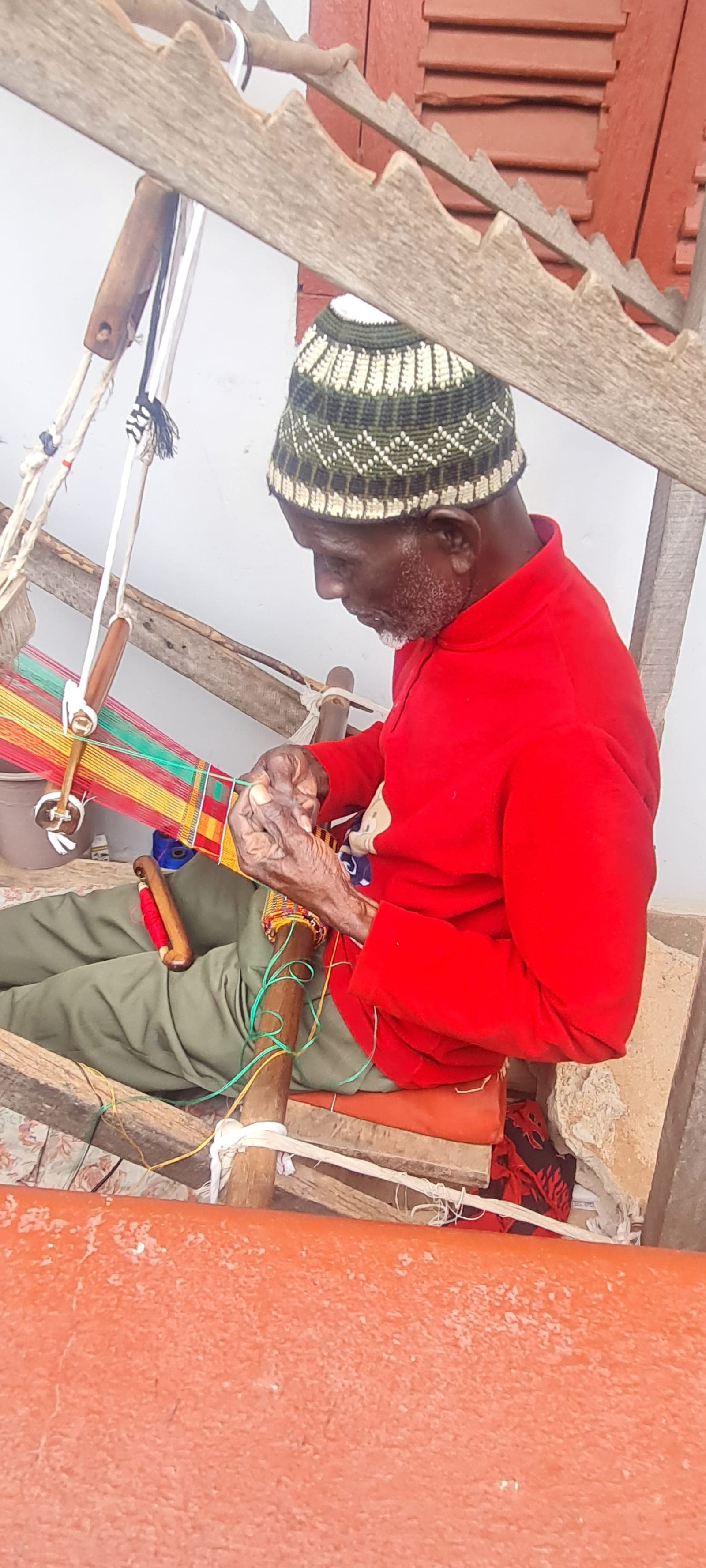 An elder weaving a red, gold and green Kente strip on a traditional wooden loom in Adanwomase
